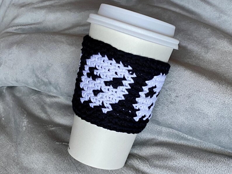 May include: A white disposable coffee cup with a white plastic lid, featuring a black and white crocheted sleeve. The sleeve displays a repeating pattern of white ghost shapes against a solid black background, wrapped around the center of the cup.