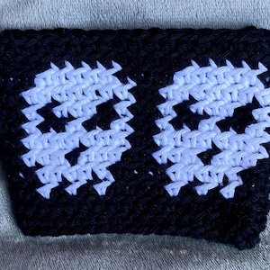 May include: A rectangular black crochet sleeve featuring two white skull motifs created with a pixelated design. The accessory is crafted from yarn with a visible textured stitch pattern, showcasing a high-contrast color scheme of black and white.