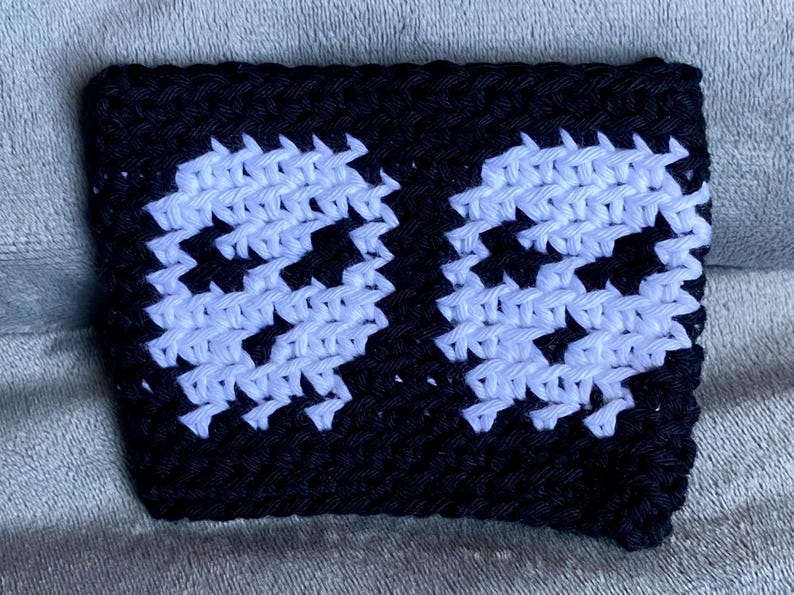 May include: A rectangular black crochet sleeve featuring two white skull motifs created with a pixelated design. The accessory is crafted from yarn with a visible textured stitch pattern, showcasing a high-contrast color scheme of black and white.