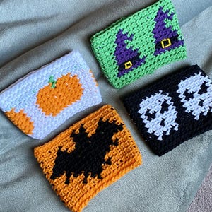 May include: Four rectangular crochet cup sleeves featuring seasonal designs. One is white with an orange pumpkin, one is green with two purple witch hats, one is black with two white skulls, and one is orange with a large black bat silhouette.