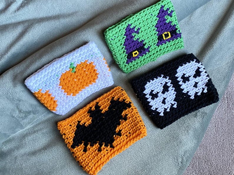 May include: Four rectangular crochet cup sleeves featuring seasonal designs. One is white with an orange pumpkin, one is green with two purple witch hats, one is black with two white skulls, and one is orange with a large black bat silhouette.