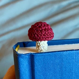May include: A small, crocheted mushroom bookmark featuring a deep red cap and a cream-colored stem. The mushroom is positioned on the top edge of a closed, bright blue hardcover book, with the stem extending downward between the pages.