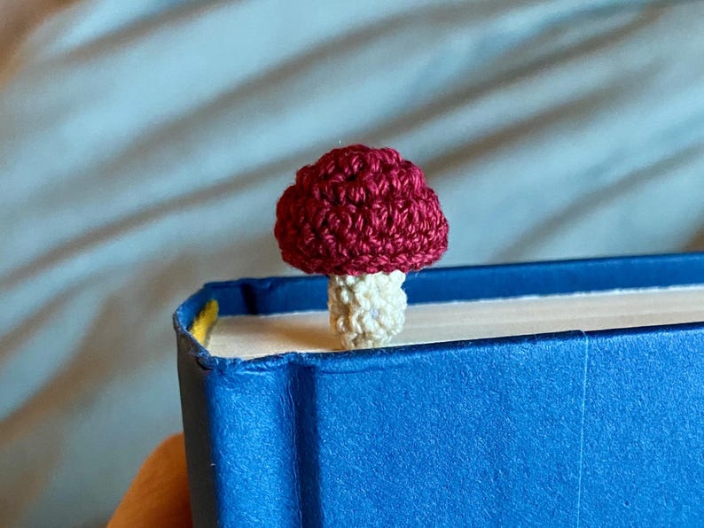 May include: A small, crocheted mushroom bookmark featuring a deep red cap and a cream-colored stem. The mushroom is positioned on the top edge of a closed, bright blue hardcover book, with the stem extending downward between the pages.