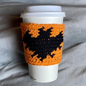 May include: A white disposable coffee cup with a white plastic lid, featuring a handmade orange crochet sleeve. The sleeve is decorated with a prominent black crocheted bat silhouette centered on the front, creating a high-contrast design.