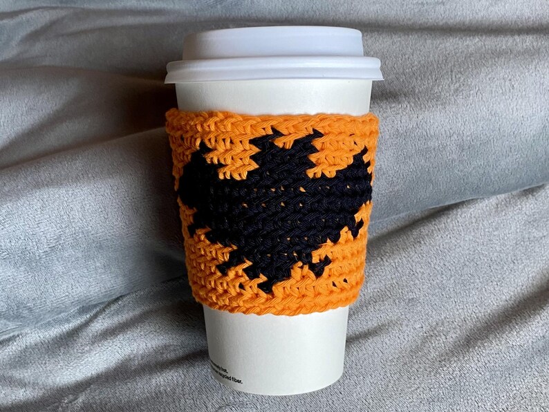 May include: A white disposable coffee cup with a white plastic lid, featuring a handmade orange crochet sleeve. The sleeve is decorated with a prominent black crocheted bat silhouette centered on the front, creating a high-contrast design.