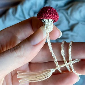 May include: A small, handcrafted crochet mushroom bookmark featuring a deep red cap and a cream-colored stem. The mushroom is attached to a long, thin, cream-colored crocheted cord that ends in a matching tassel, designed for marking pages in a book.