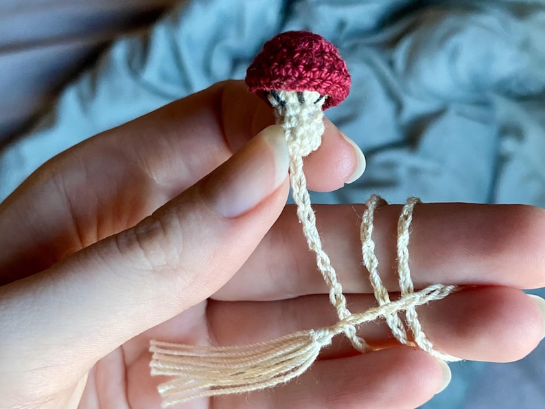 May include: A small, handcrafted crochet mushroom bookmark featuring a deep red cap and a cream-colored stem. The mushroom is attached to a long, thin, cream-colored crocheted cord that ends in a matching tassel, designed for marking pages in a book.