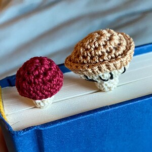 May include: Two small, crocheted mushroom-shaped bookmarks resting on the top edge of a closed blue book. One mushroom has a deep red cap with a cream-colored stem, while the other features a light brown cap with a cream-colored stem.