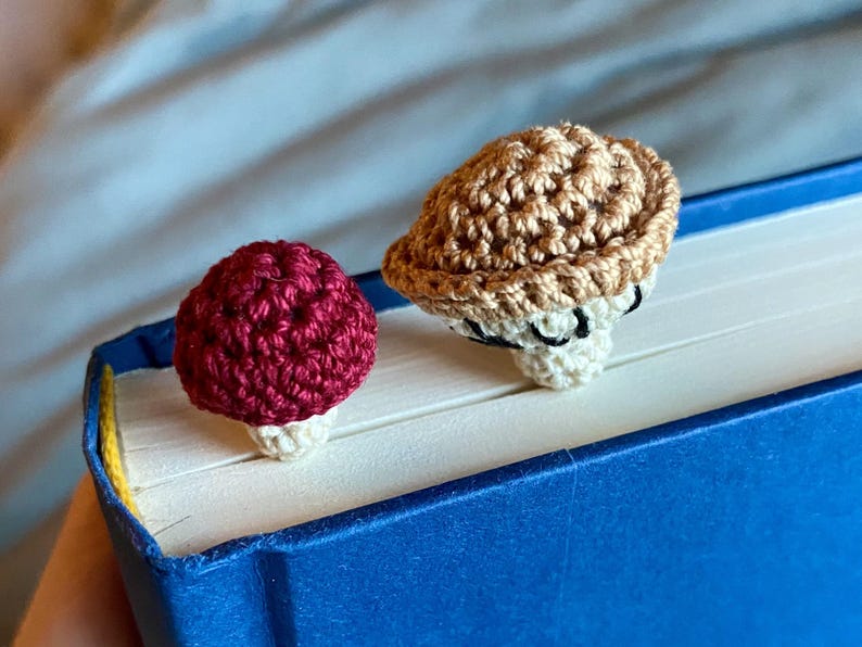 May include: Two small, crocheted mushroom-shaped bookmarks resting on the top edge of a closed blue book. One mushroom has a deep red cap with a cream-colored stem, while the other features a light brown cap with a cream-colored stem.