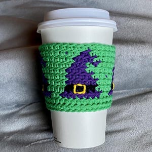 May include: A white disposable coffee cup featuring a lime green crocheted sleeve. The sleeve displays a repeating pattern of purple witch hats with black bands and yellow buckles, crafted with a textured knit stitch design.