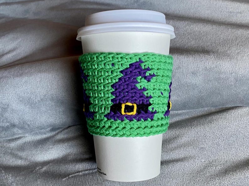 May include: A white disposable coffee cup featuring a lime green crocheted sleeve. The sleeve displays a repeating pattern of purple witch hats with black bands and yellow buckles, crafted with a textured knit stitch design.