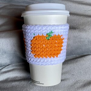 May include: A white disposable coffee cup with a lid, featuring a handmade crochet sleeve. The sleeve is light purple with a prominent orange pumpkin design in the center, accented by a small green stem detail on the top of the pumpkin.