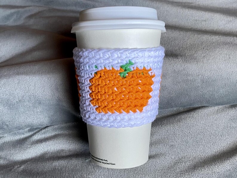 May include: A white disposable coffee cup with a lid, featuring a handmade crochet sleeve. The sleeve is light purple with a prominent orange pumpkin design in the center, accented by a small green stem detail on the top of the pumpkin.