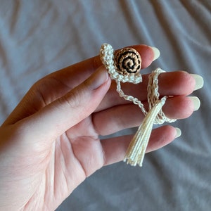 Puede incluir: Un amuleto de caracol de ganchillo con una concha marr&oacute;n y negra y un borla blanca.