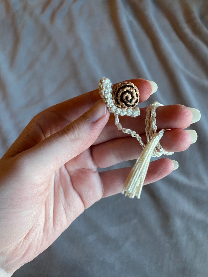 Puede incluir: Un amuleto de caracol de ganchillo con una concha marr&oacute;n y negra y un borla blanca.