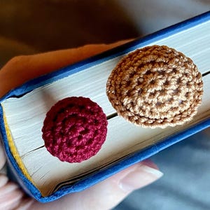 May include: Two round, crocheted yarn toppers attached to a blue book. One topper is a deep burgundy red, and the other is a light tan brown. Both items feature a textured, circular stitch pattern and are positioned on the top edge of the book.