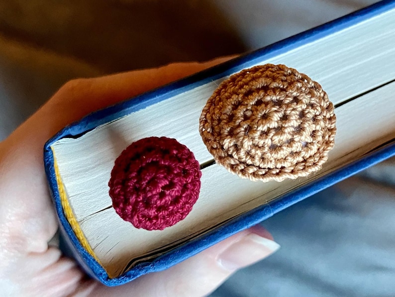 May include: Two round, crocheted yarn toppers attached to a blue book. One topper is a deep burgundy red, and the other is a light tan brown. Both items feature a textured, circular stitch pattern and are positioned on the top edge of the book.