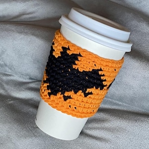 May include: A white disposable coffee cup featuring a bright orange crocheted sleeve. The sleeve is decorated with a prominent black crocheted bat silhouette centered on the front, providing a textured contrast against the vibrant orange yarn background.