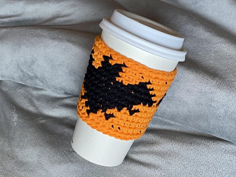 May include: A white disposable coffee cup featuring a bright orange crocheted sleeve. The sleeve is decorated with a prominent black crocheted bat silhouette centered on the front, providing a textured contrast against the vibrant orange yarn background.