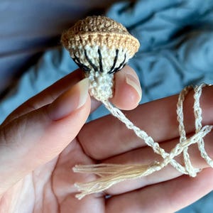 May include: A small, crocheted mushroom ornament featuring a tan cap with a cream-colored underside and dark brown stitched gills. The mushroom is attached to a long, thin, cream-colored braided cord that ends in a small, matching tassel.