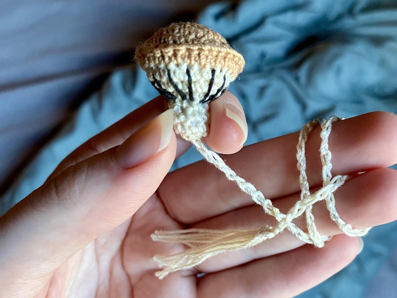 May include: A small, crocheted mushroom ornament featuring a tan cap with a cream-colored underside and dark brown stitched gills. The mushroom is attached to a long, thin, cream-colored braided cord that ends in a small, matching tassel.