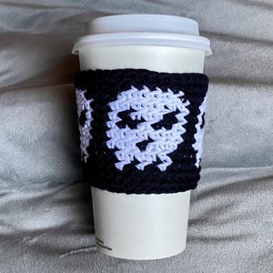 May include: A white disposable coffee cup featuring a black and white crocheted sleeve. The sleeve displays a repeating pattern of white skull motifs against a solid black background, crafted with a visible yarn texture and a secure fit.
