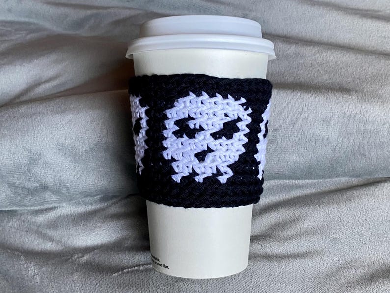 May include: A white disposable coffee cup featuring a black and white crocheted sleeve. The sleeve displays a repeating pattern of white skull motifs against a solid black background, crafted with a visible yarn texture and a secure fit.