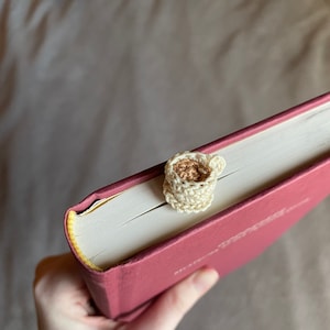 Top view of a crocheted teacup bookmark sticking out of the top of a red hardcover book. The book is held by hand in front of a gray cloth background. The teacup is off white and has a saucer. The light brown tea inside is visible from this angle.