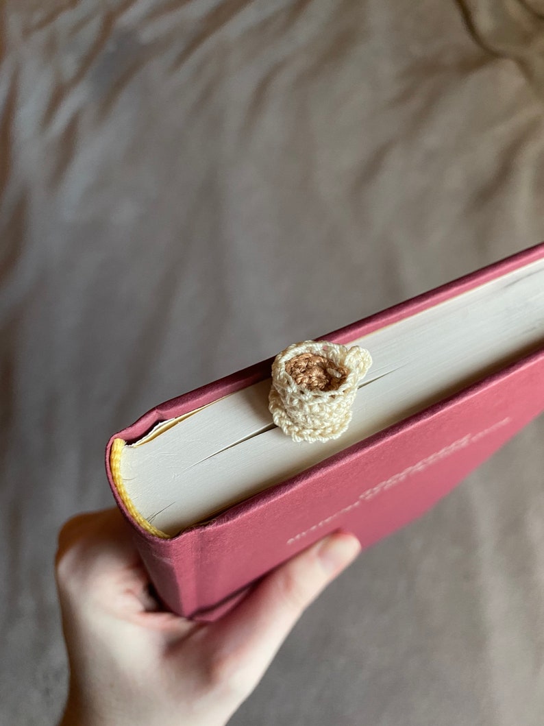 Top view of a crocheted teacup bookmark sticking out of the top of a red hardcover book. The book is held by hand in front of a gray cloth background. The teacup is off white and has a saucer. The light brown tea inside is visible from this angle.