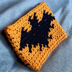 May include: A square crochet coaster featuring a vibrant orange background with a centered black bat silhouette. The piece is crafted with a visible yarn texture and consistent stitch pattern, showcasing a high-contrast design in orange and black.