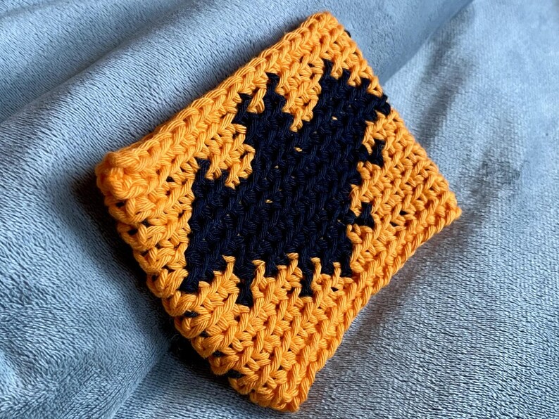 May include: A square crochet coaster featuring a vibrant orange background with a centered black bat silhouette. The piece is crafted with a visible yarn texture and consistent stitch pattern, showcasing a high-contrast design in orange and black.