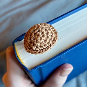 May include: A small, round, light brown crocheted accessory with a textured, knotted surface. The item is attached to the top edge of a closed book with a solid blue cover, resting against the white pages of the book's spine and top edge.