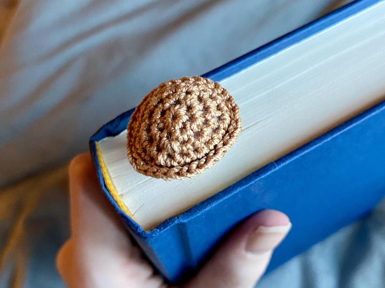 May include: A small, round, light brown crocheted accessory with a textured, knotted surface. The item is attached to the top edge of a closed book with a solid blue cover, resting against the white pages of the book's spine and top edge.