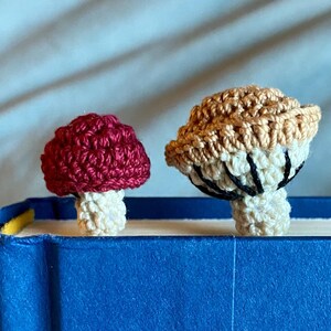 May include: Two crochet mushroom bookmarks resting on the top edge of a blue book. The smaller mushroom has a deep red cap and a cream stem. The larger mushroom features a tan cap with visible cream-colored gills and black stitching details.