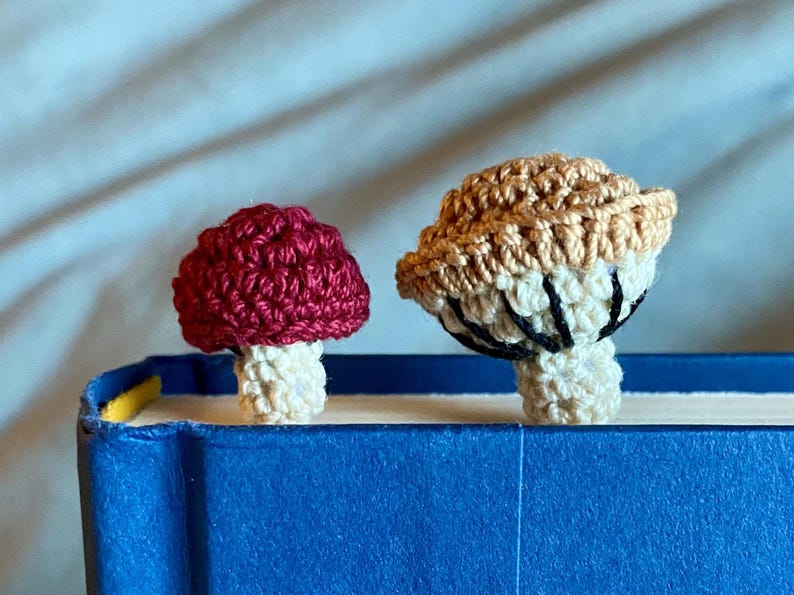 May include: Two crochet mushroom bookmarks resting on the top edge of a blue book. The smaller mushroom has a deep red cap and a cream stem. The larger mushroom features a tan cap with visible cream-colored gills and black stitching details.