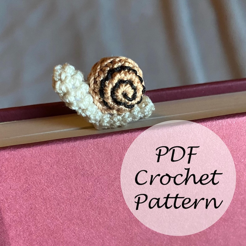 Puede incluir: Un marcap&aacute;ginas de ganchillo en forma de caracol con una concha marr&oacute;n y negra. El caracol est&aacute; sentado en un libro rosa con el texto "PDF Crochet Pattern" en un c&iacute;rculo blanco.