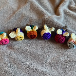 May include: Seven crocheted bee keychains in various colors, including pink, yellow, orange, purple, red, blue, and gray. Each bee has white wings and a black face.