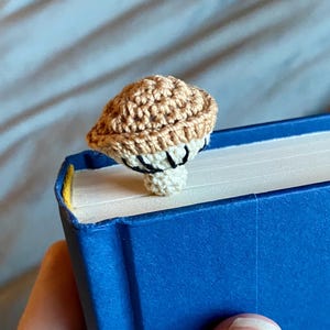 May include: A crochet mushroom bookmark featuring a tan cap with a textured surface and a cream-colored stem. The underside of the mushroom cap displays black stitched gills. The bookmark is positioned on the top edge of a blue hardcover book.