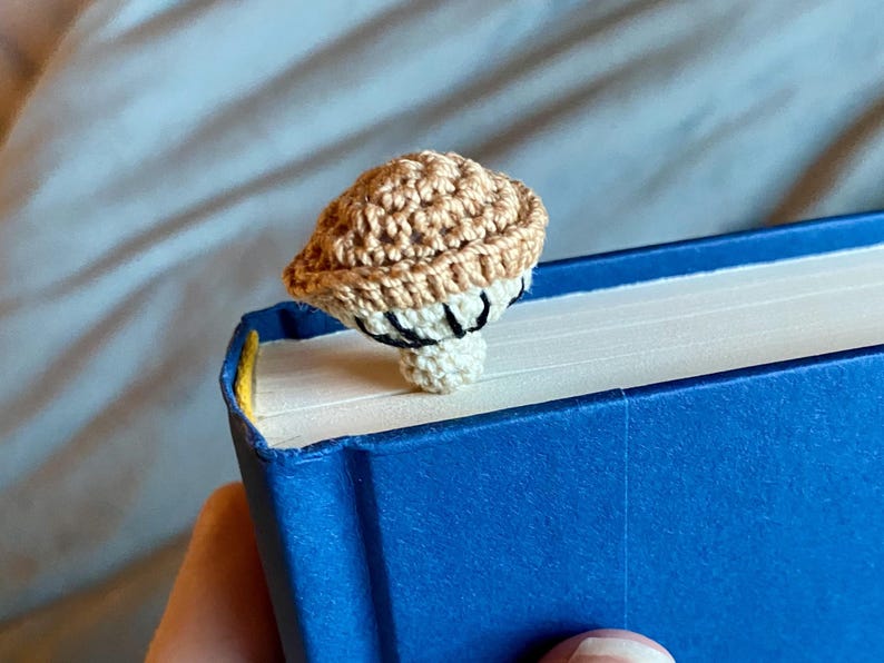 May include: A crochet mushroom bookmark featuring a tan cap with a textured surface and a cream-colored stem. The underside of the mushroom cap displays black stitched gills. The bookmark is positioned on the top edge of a blue hardcover book.