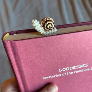 Puede incluir: Un marcap&aacute;ginas de ganchillo en forma de caracol en colores marr&oacute;n, blanco y negro. El caracol est&aacute; encima de un libro cerrado con el t&iacute;tulo "Goddesses Mysteries of the Feminine" en el lomo.