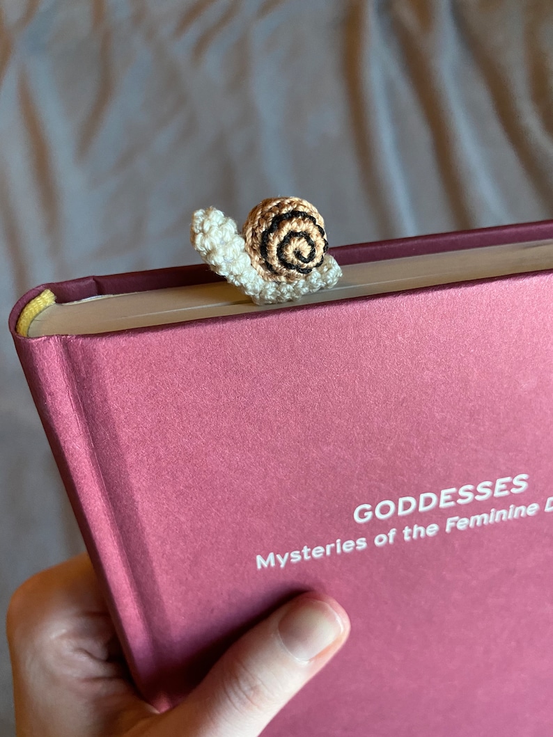 Puede incluir: Un marcap&aacute;ginas de ganchillo en forma de caracol en colores marr&oacute;n, blanco y negro. El caracol est&aacute; encima de un libro cerrado con el t&iacute;tulo "Goddesses Mysteries of the Feminine" en el lomo.