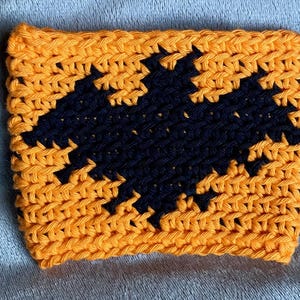 May include: A rectangular crochet cup cozy featuring a vibrant orange background with a prominent black bat silhouette centered in the middle. The piece is crafted with a visible yarn stitch pattern, creating a textured surface throughout the design.