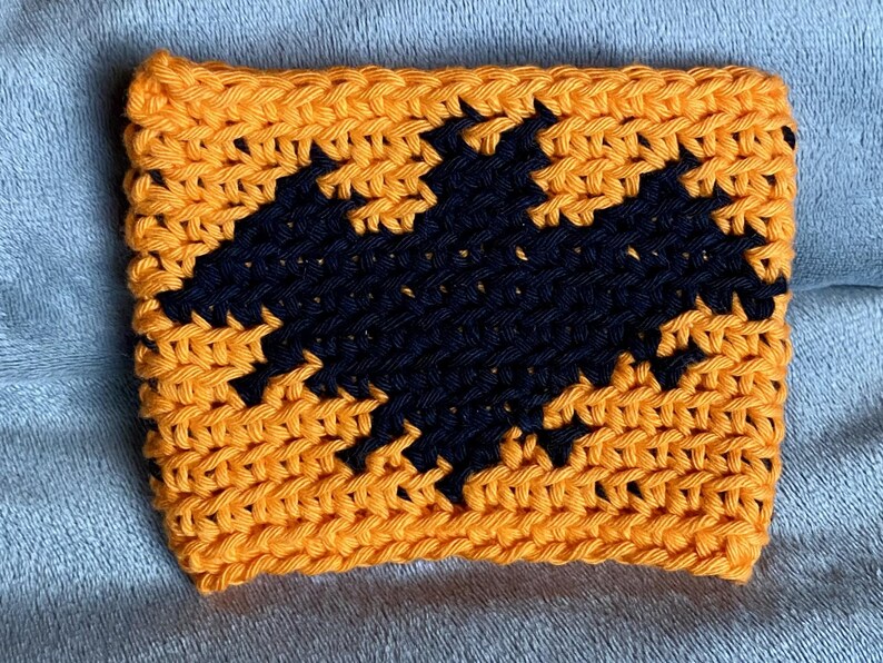 May include: A rectangular crochet cup cozy featuring a vibrant orange background with a prominent black bat silhouette centered in the middle. The piece is crafted with a visible yarn stitch pattern, creating a textured surface throughout the design.