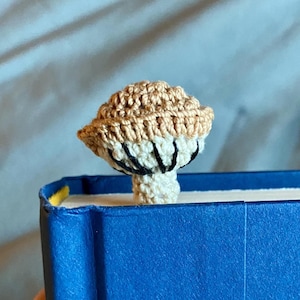 May include: A crochet mushroom bookmark featuring a tan cap and a cream-colored stem. The underside of the mushroom cap displays visible black stitched gills. The mushroom is positioned upright, emerging from the top edge of a bright blue book.
