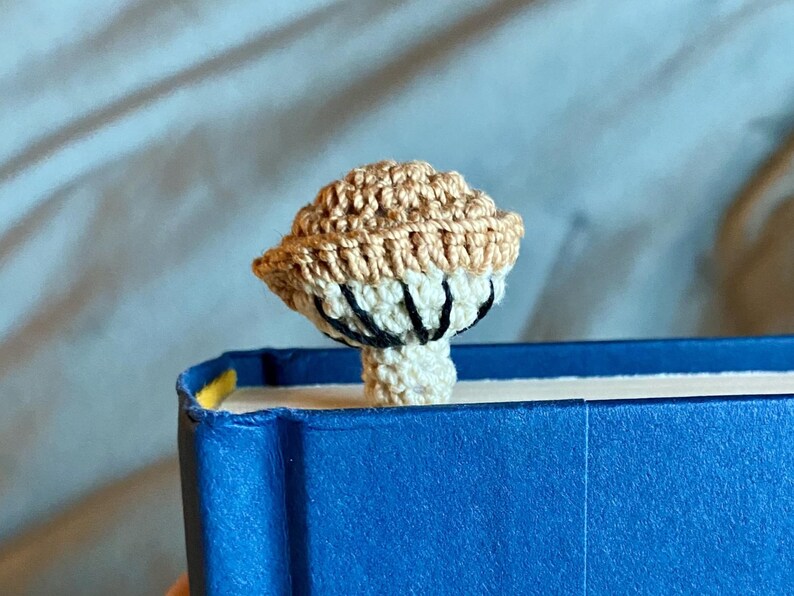 May include: A crochet mushroom bookmark featuring a tan cap and a cream-colored stem. The underside of the mushroom cap displays visible black stitched gills. The mushroom is positioned upright, emerging from the top edge of a bright blue book.