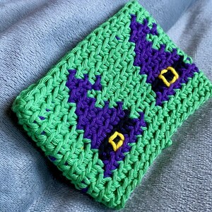 May include: A rectangular crochet pouch featuring a bright green base with two dark purple witch hat motifs. Each purple hat is accented with a small, embroidered yellow square buckle detail. The item is constructed with a visible, textured yarn stitch.