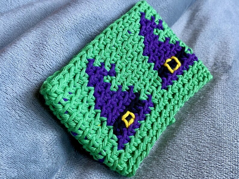 May include: A rectangular crochet pouch featuring a bright green base with two dark purple witch hat motifs. Each purple hat is accented with a small, embroidered yellow square buckle detail. The item is constructed with a visible, textured yarn stitch.