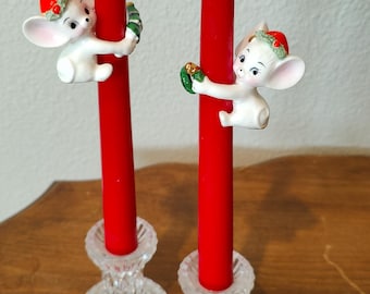 May include: Two red taper candles held by white ceramic mouse figurines wearing red hats with green wreaths. Each candle sits in a clear, ribbed glass candlestick holder. The mice are positioned to appear as if they are climbing the candles.