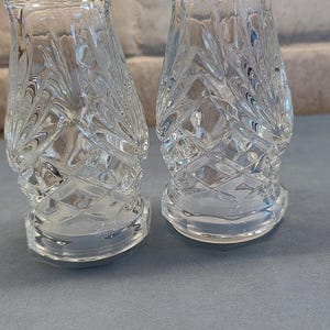May include: Two clear glass salt and pepper shakers with a textured, leaf-like pattern. The shakers have a flared base and a decorative, scalloped top. They are displayed on a light blue surface.
