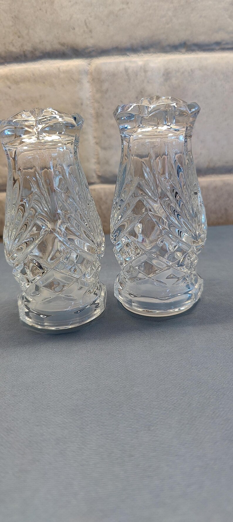 May include: Two clear glass salt and pepper shakers with a textured, leaf-like pattern. The shakers have a flared base and a decorative, scalloped top. They are displayed on a light blue surface.